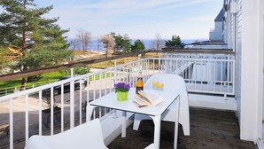 Ein Balkon mit weißen Möbeln, einem Tisch mit Buch und Getränken sowie Blick auf das Meer und Bäume.