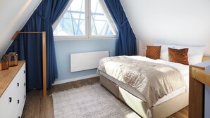 Ein Schlafzimmer mit schrägen Wänden, einem großen Fenster mit blauen Vorhängen, einem Bett mit gemustertem Überwurf und einem Nachttisch mit Lampe.