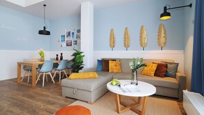 Ein modernes Wohnzimmer mit grauem Sofa, orangefarbenem Hocker und einem runden Couchtisch. Es gibt einen Essbereich mit Holztisch und Stühlen. Die Wände sind blau mit Blättermustern und es gibt eine Deckenleuchte.