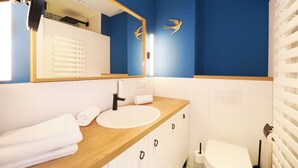 Une salle de bain moderne avec un mur d’accent bleu, un meuble-lavabo en bois, un lavabo blanc, des toilettes et un miroir orné d’un motif d’oiseau doré.