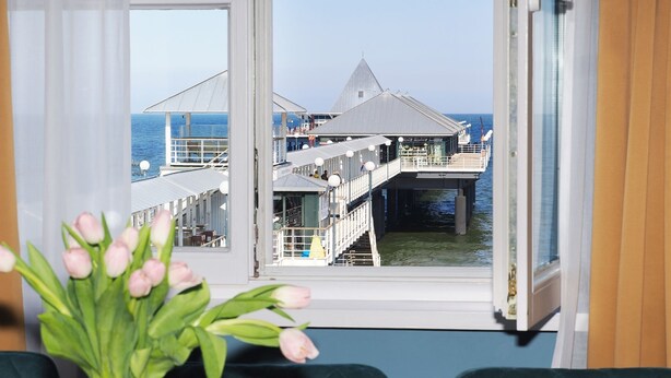 Blick aus einem Fenster mit Balkon auf einen Steg und das Meer.