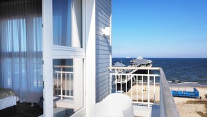 Ein Balkon mit Blick auf Strand und Meer, weiße Gartenmöbel und ein Gebäude mit Glastür.
