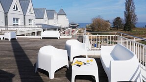 Een houten terras met witte kunststof meubels, uitzicht op een gebouw met een grijs dak en een heldere lucht.