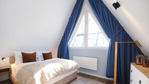 Ein Schlafzimmer mit Bett, Nachttisch, Kommode, einem Fenster mit Vorhängen und einer Heizung.