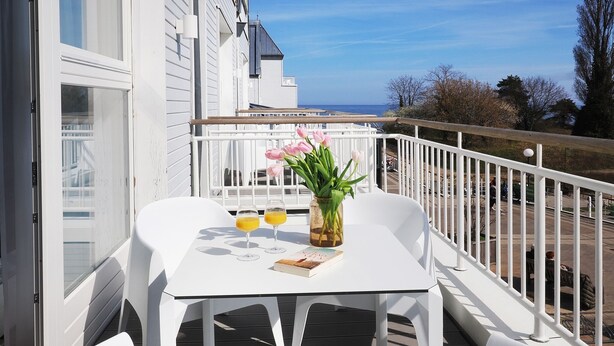 Una mesa blanca con dos vasos de zumo de naranja, un libro y un jarrón con flores sobre una terraza de madera.