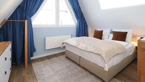Un dormitorio con techo inclinado, cama con edredón blanco y almohadas marrones, mesita de noche de madera, radiador blanco y ventana con cortinas azules.