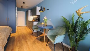 A modern bedroom with a wooden floor, a bed with a patterned bedspread, a small kitchenette, a dining area with a table and chairs, and a wall with a decorative bird and the phrase "Life is". - 1-room vacation apartment with balcony (Seebad Heringsdorf)