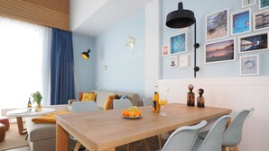 Een moderne woonkamer met een houten eettafel, lichtblauwe stoelen en een bank met kussens. Er is een wandlamp, een verzameling ingelijste foto’s en een fles sinaasappelsap op de tafel.