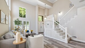 A spacious living room with a high ceiling, a staircase with white railings, and a large window providing natural light. - 5BR Home Modern & Spacious for Business & Family (San Jose)