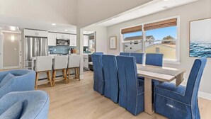 A modern kitchen with white cabinets, stainless steel appliances, and a dining area with blue upholstered chairs and a wooden table. - Fenwick Shores House II (Selbyville)