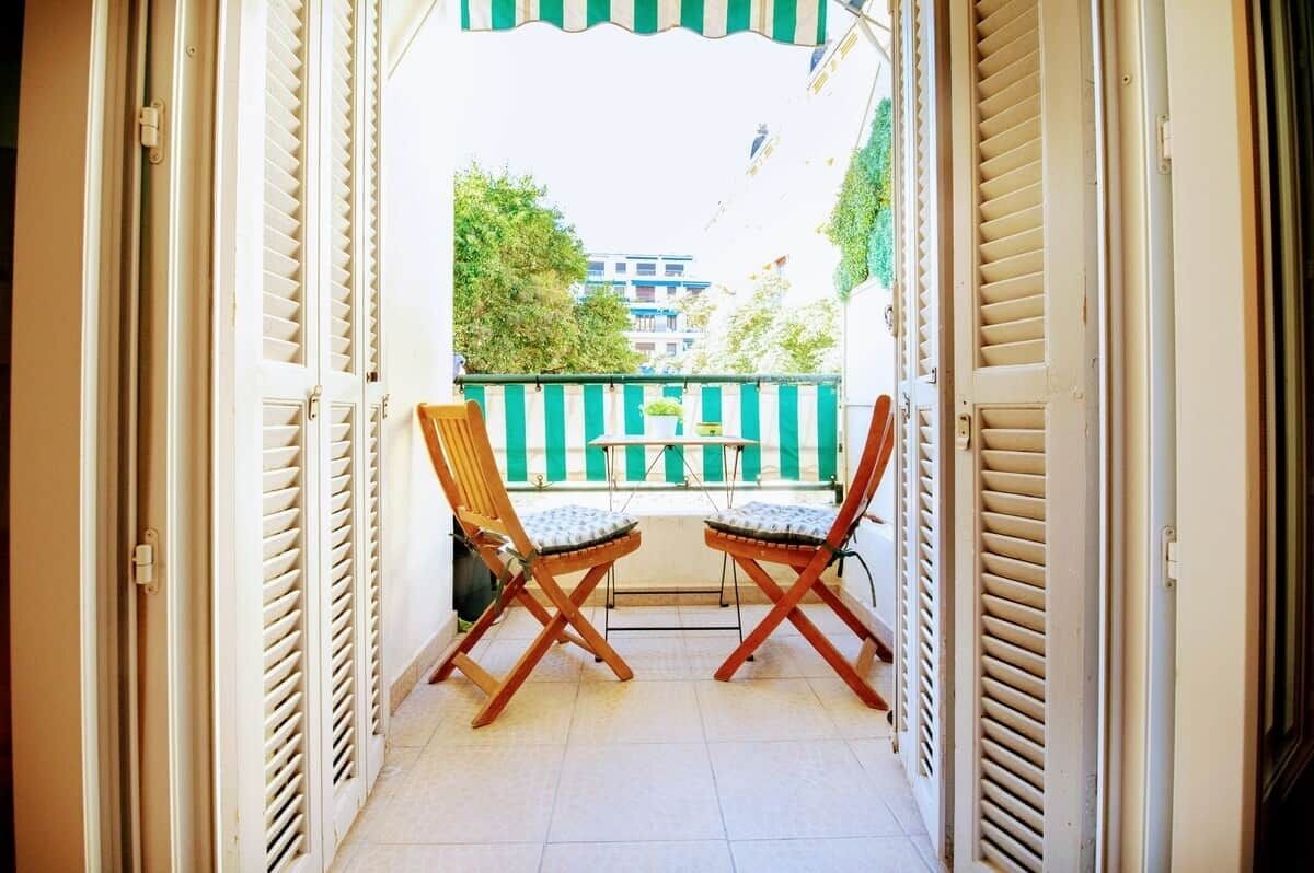 Een open balkon met twee houten stoelen en een kleine tafel, een groen gestreept hek en gebouwen op de achtergrond.