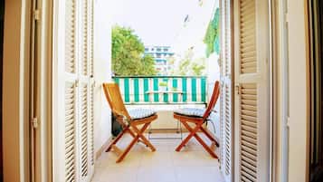 Een open balkon met twee houten stoelen en een kleine tafel, een groen gestreept hek en gebouwen op de achtergrond.