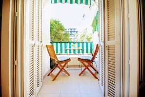 An open balcony with two wooden chairs and a small table, a green-striped fence, and buildings in the background.