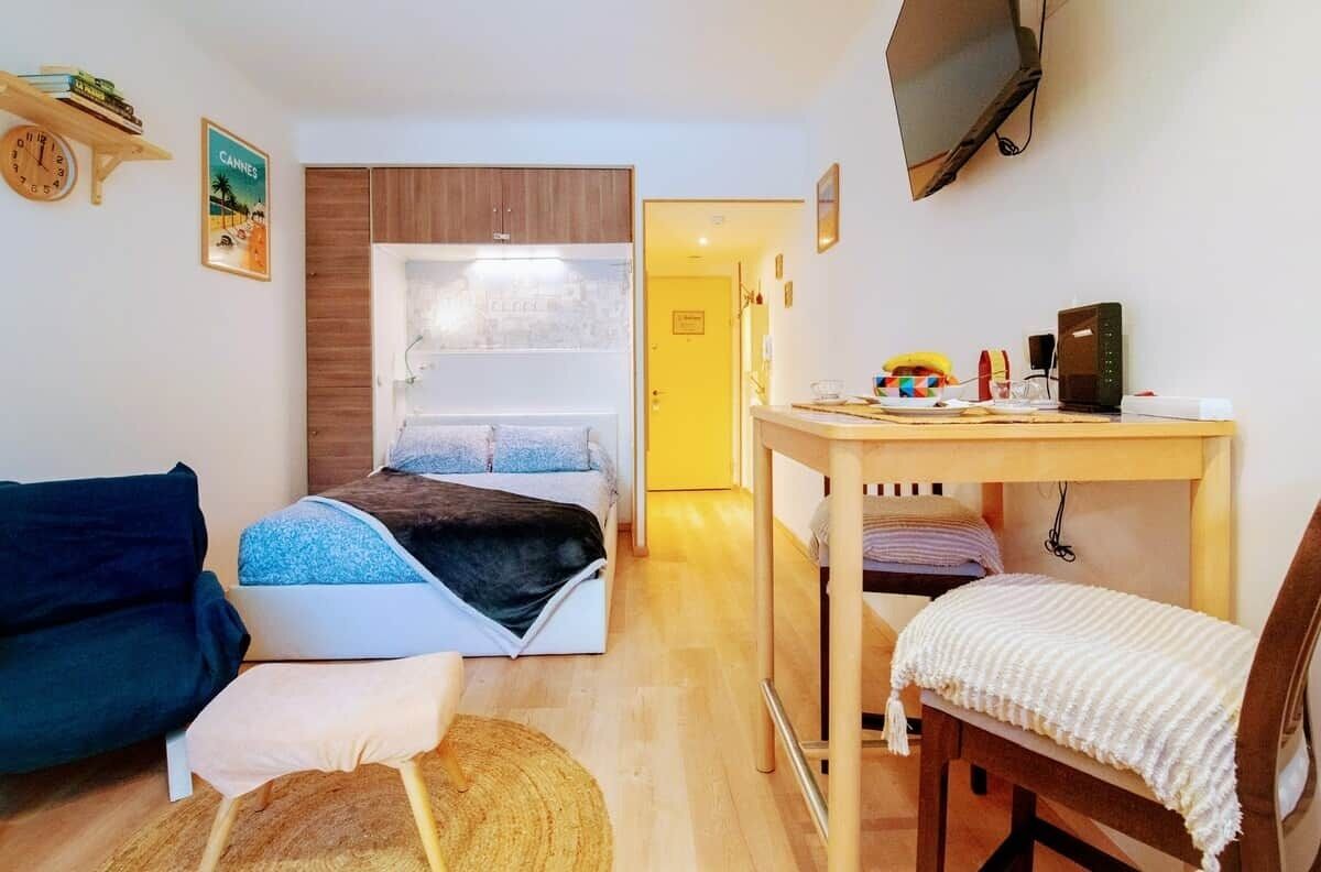 A compact room with a bed, a small table, a chair, a sofa, and a television mounted on the wall.