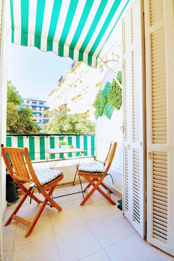 A balcony with two wooden chairs and a small table, a green and white striped awning, and a view of buildings and greenery.