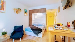 Ein Schlafzimmer mit Bett, Sessel, Schreibtisch mit Computer und Wandfernseher.