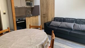 A compact living space with a dining table, a black leather sofa, a kitchen area with a microwave, and wooden cabinets.