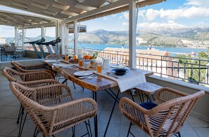 Une terrasse avec des chaises en osier, une table dressée pour le repas et une vue panoramique sur les montagnes et l’eau.