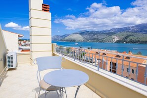 Un balcon avec une table ronde et deux chaises, donnant sur une étendue d’eau et sur des bâtiments aux toits recouverts de tuiles rouges.