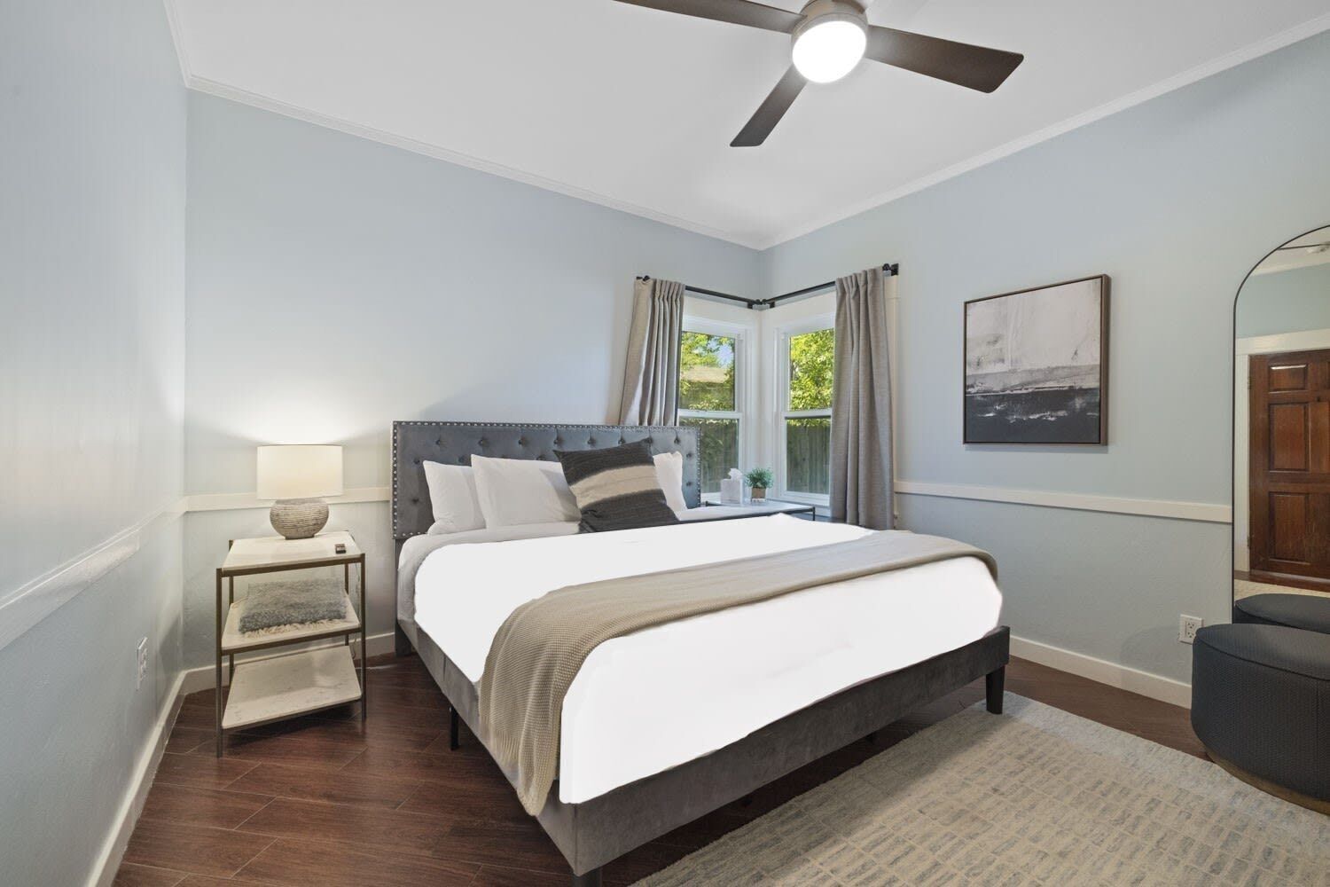 A bedroom with a large bed, a bedside table with a lamp, a ceiling fan, and a window with curtains.