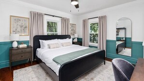 Un dormitorio con cama, mesitas de noche, ventilador de techo, ventanas con cortinas, un espejo y una silla.
