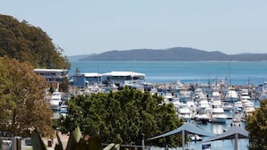 A marina with numerous boats docked, a building with a sign reading "FISH & CHIPS", and a hilly landscape in the background. - The Waterview, 2 76 Magnus St -  complete style, comfort, luxury and spectacular waterviews (Nelson Bay)
