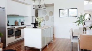 A modern kitchen with white cabinetry, a central island, and a dining area with a table and chairs. - The Waterview, 2 76 Magnus St -  complete style, comfort, luxury and spectacular waterviews (Nelson Bay)