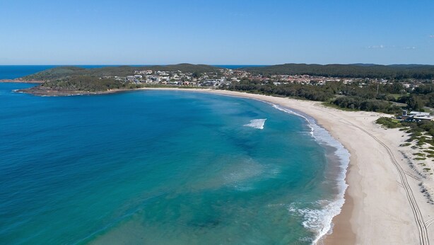 Vista aérea de una zona costera con playa de arena, aguas azules cristalinas y un pueblo con edificios al fondo.