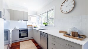 Una cocina moderna con armarios blancos, horno empotrado, lavavajillas y un amplio ventanal que proporciona luz natural.
