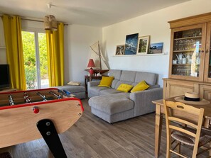 Una sala de estar con sofá, mesa de centro de madera, mesa de comedor con sillas, mesa de fútbol de mesa y un mueble con puertas de vidrio.