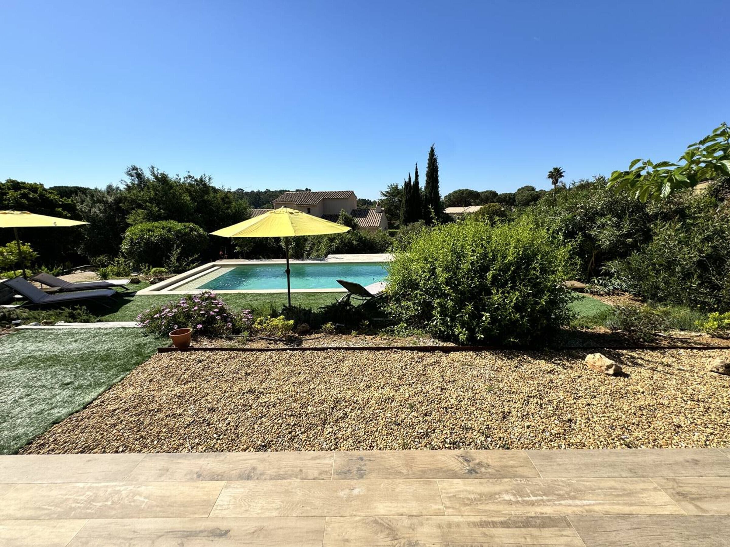 A swimming pool with a tiled deck, surrounded by greenery and a clear sky.