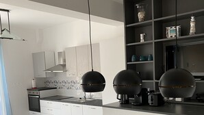 Et moderne køkken med hvide skabe, en mørk bordplade og hængende lamper. Der er et spiseområde med et træbord og stole.