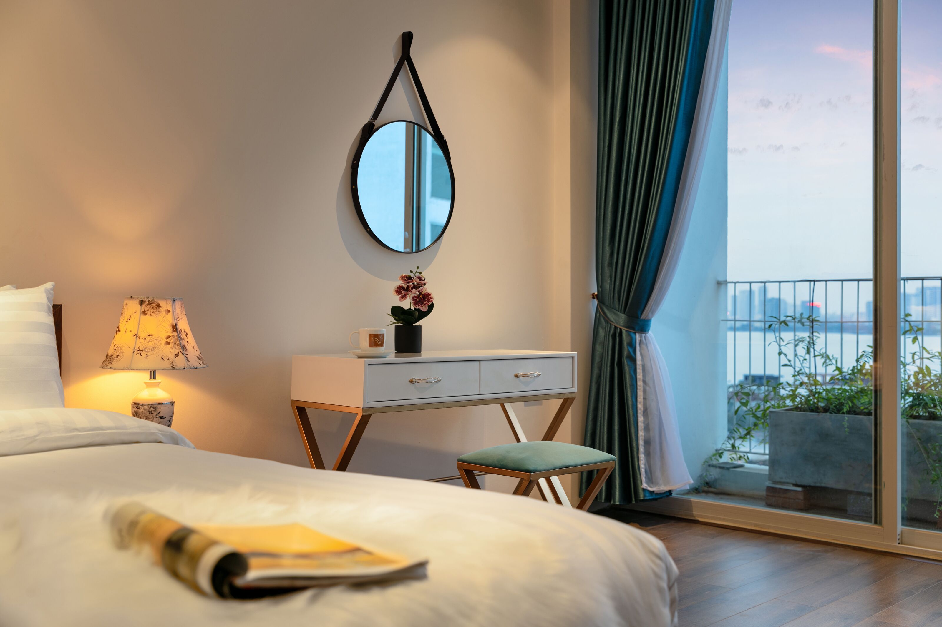 A hotel room with a bed, a desk with a lamp, a small stool, a mirror, and a view of the outside balcony.
