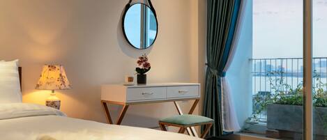 A hotel room with a bed, a desk with a lamp, a small stool, a mirror, and a view of the outside balcony.