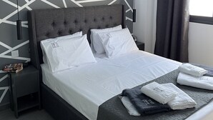 Moderni hotellihuone, jossa on geometrisesti kuvioitu seinä, harmaa verhoiltu sänky, valkoiset vuodevaatteet sekä yöpöytä, jonka päällä on kirja ja lamppu.