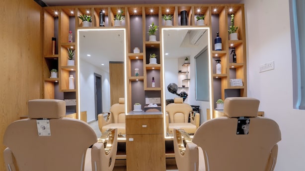 A salon with two beige chairs, wooden shelves with plants and toiletries, and a mirror reflecting the room. - The Royal Bihar (Danapur)