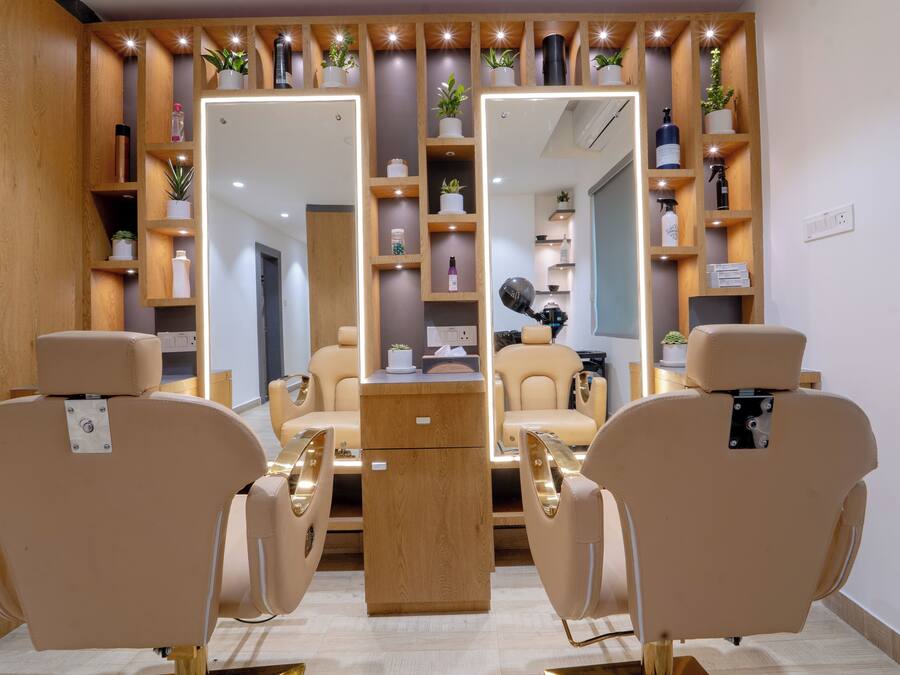 Een kapsalon met twee beige stoelen, houten planken met planten en toiletartikelen, en een spiegel die de ruimte weerspiegelt.