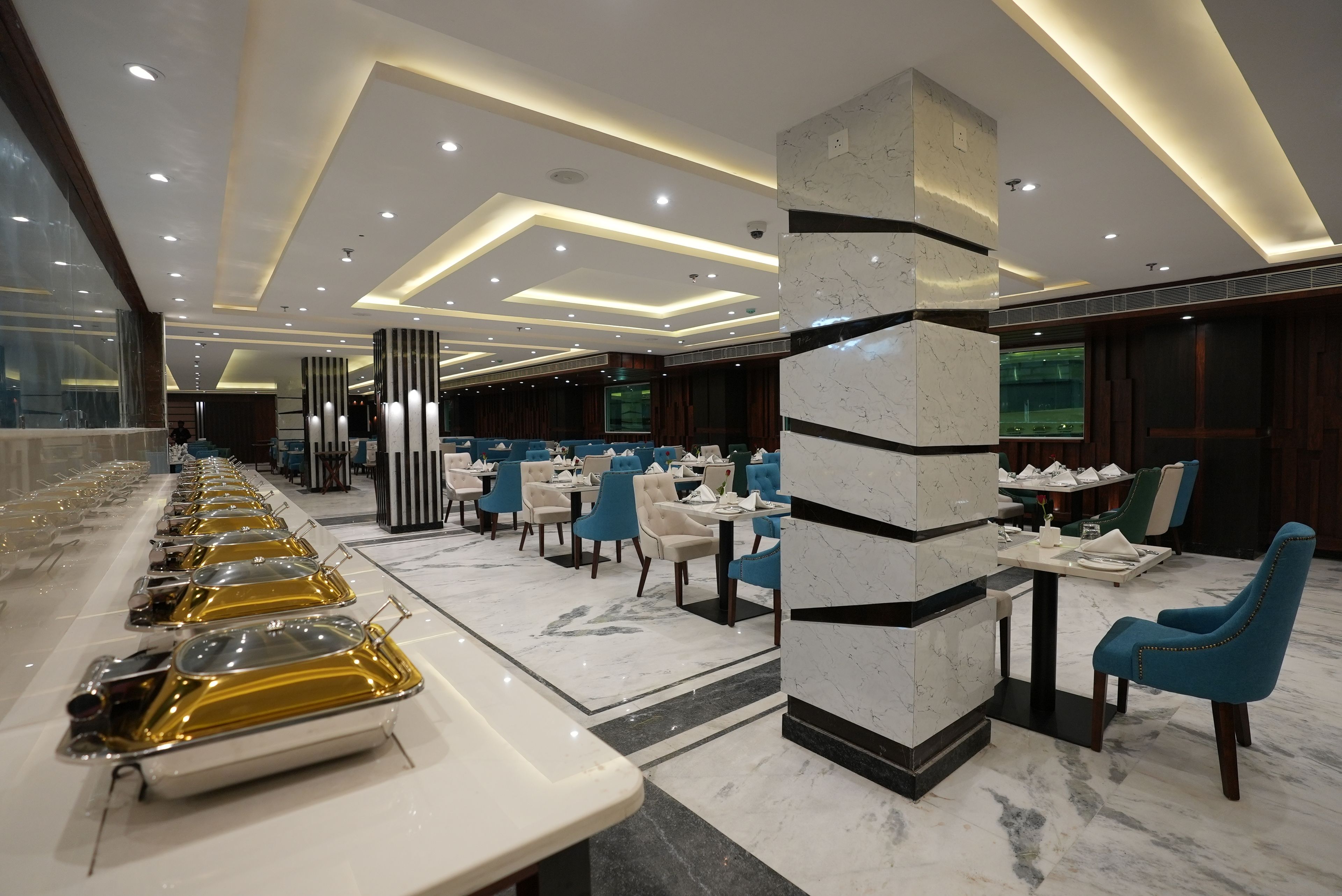 Un comedor de hotel con varias mesas y sillas, una larga barra de buffet y un diseño de techo moderno.