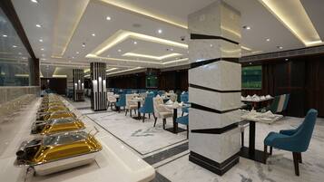 Un comedor de hotel con varias mesas y sillas, una larga barra de buffet y un diseño de techo moderno.