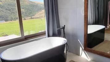 A modern bathroom with a freestanding bathtub, a large mirror, and a view of a green landscape through a window.