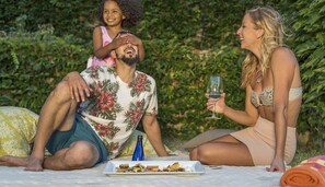 Una familia de tres sobre una manta de picnic, con comida y bebidas, rodeada de follaje.