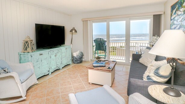 A living room with a television, a coffee table, and a sofa. There is a balcony with a view of the ocean. - Tradewinds 63 - 3rd Floor Ocean View Unit with Stunning View (St. Augustine)