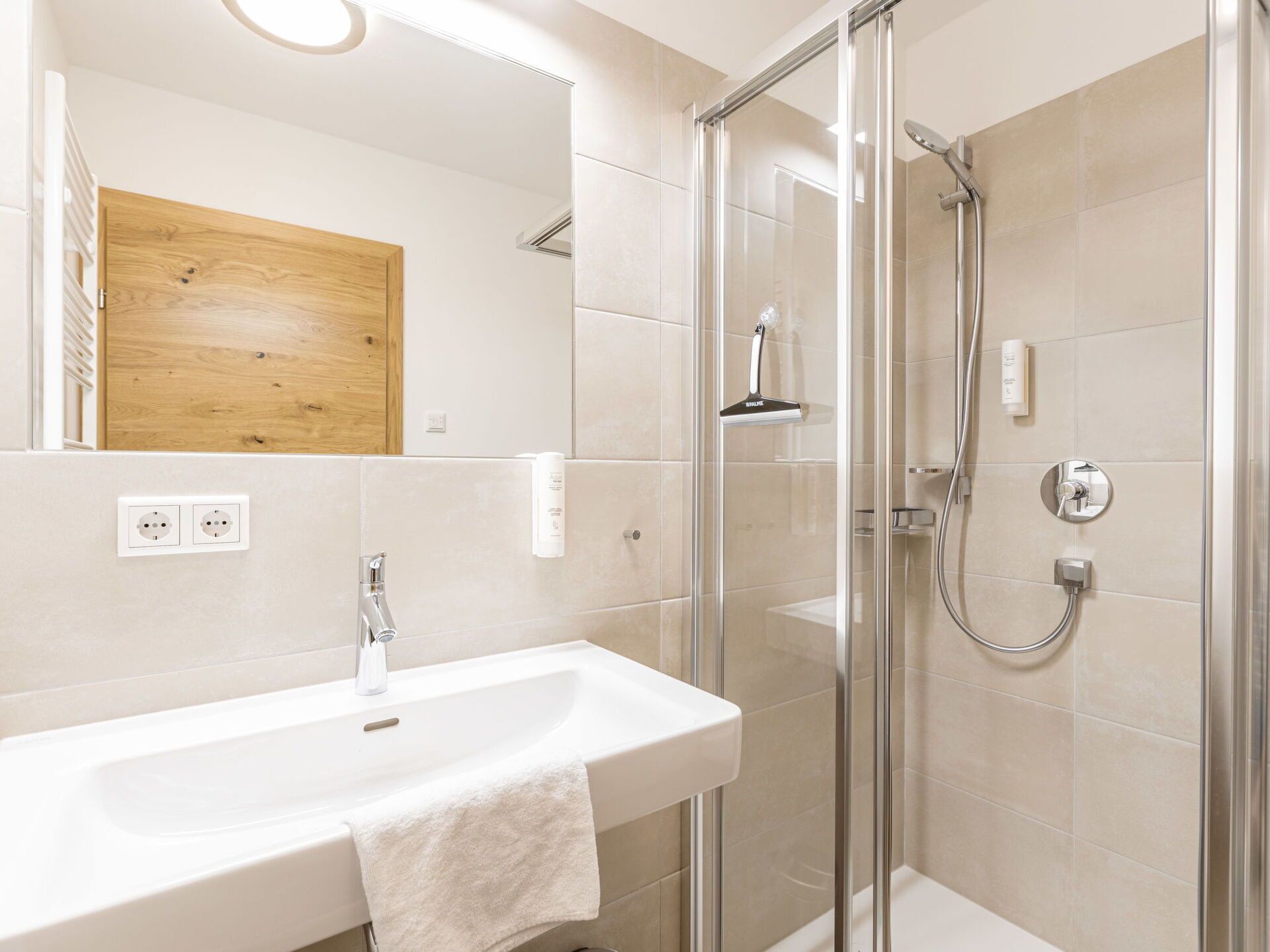 Ein modernes Badezimmer mit einer gläsernen Duschkabine, einem weißen Waschbecken und einer Holztür.