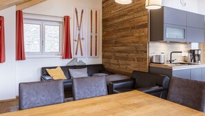 Un soggiorno moderno con soffitto in legno, un divano, un tavolo da pranzo e un'area cucina con mobili grigi e una macchina da caffè in acciaio inossidabile.