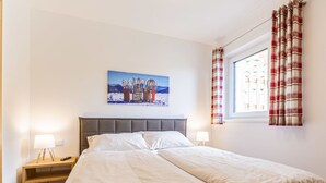 A neatly made double bed with a grey headboard, two bedside tables with lamps, a window with red and white curtains, and a painting of a mountain landscape on the wall. - Lovely apartment for 8 people with WIFI, TV and terrace (Brandstätter)