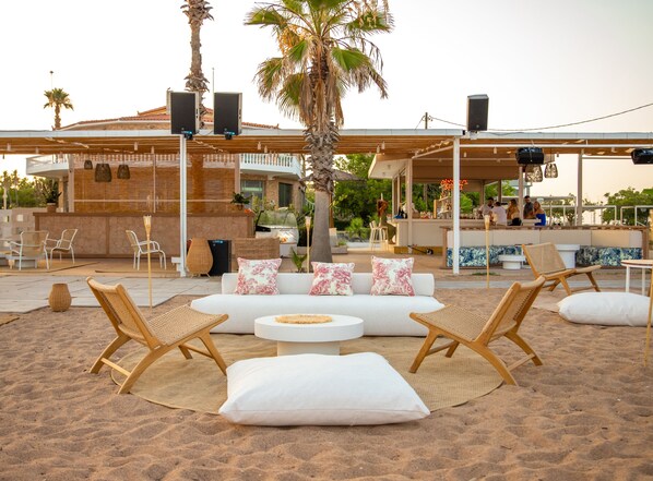 A beachside lounge with a white sofa set, wicker chairs, and a bar area with patrons.