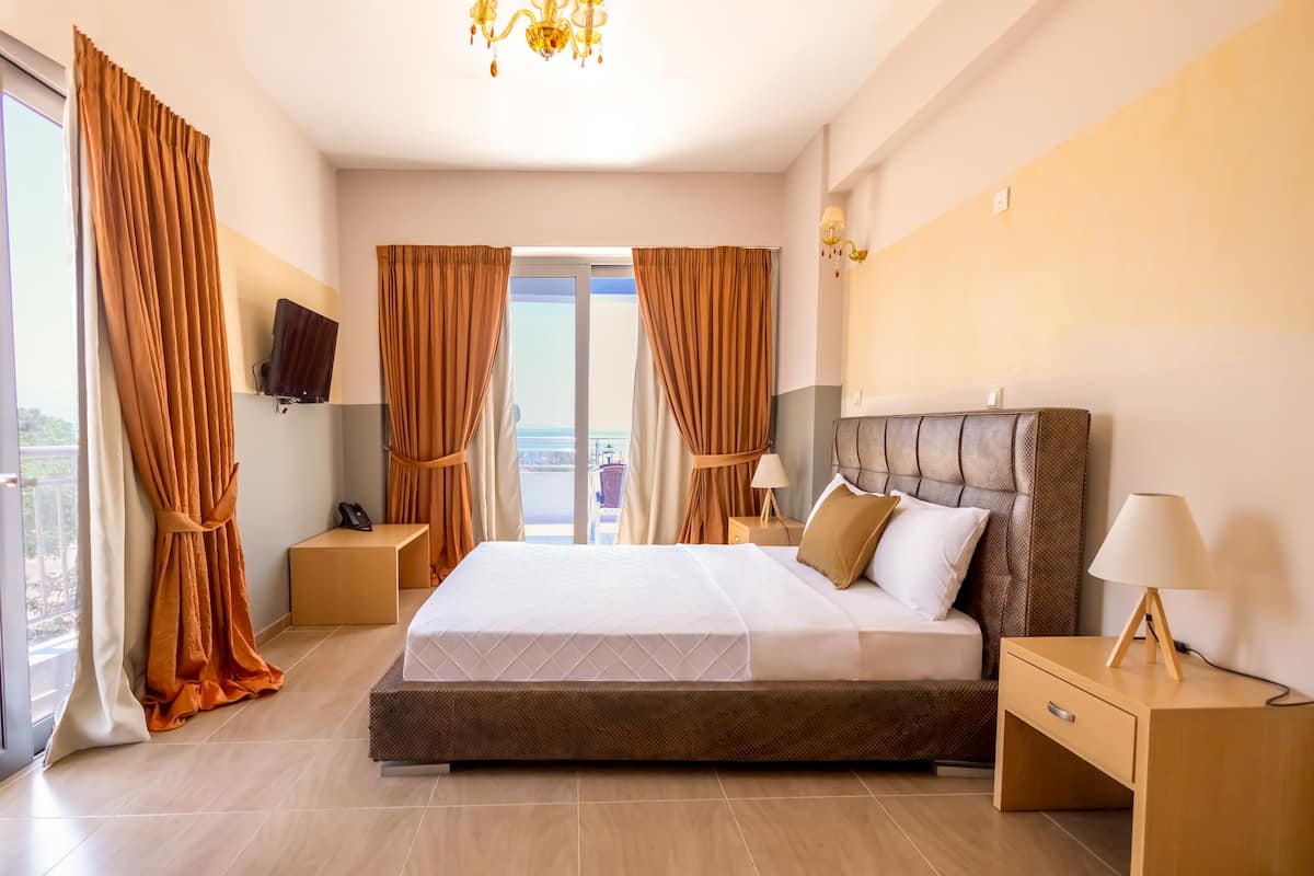 A modern hotel room with a large bed, a flat-screen TV mounted on the wall, a bedside table with a lamp, and a view of the city through the window.