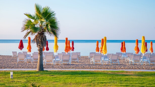 A beach with rows of white lounge chairs and colorful umbrellas, a palm tree, and a clear view of the sea. - Buka Sandy Beach  (Messini)