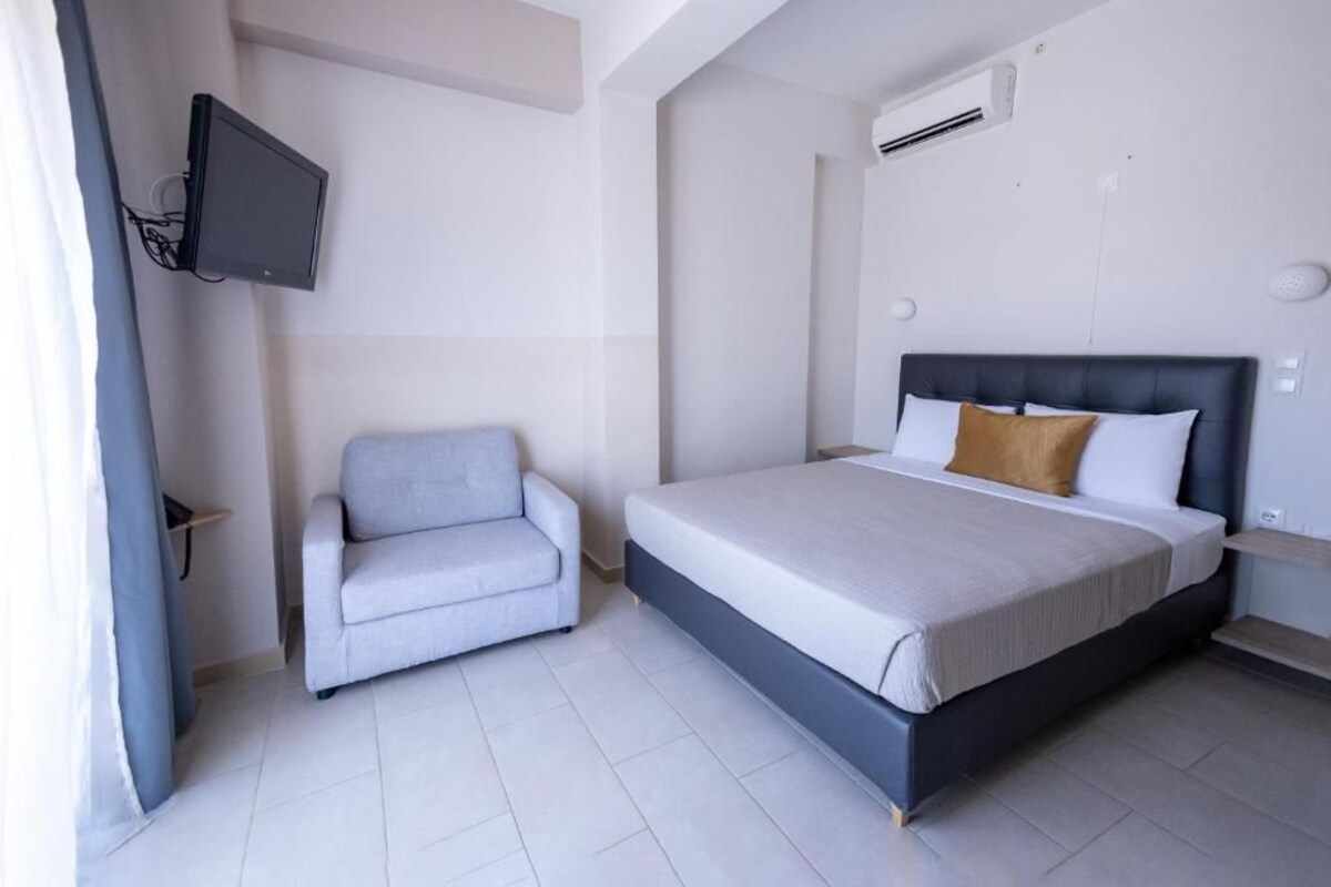 A bedroom with a bed, a grey armchair, a mounted TV, and an air conditioning unit.