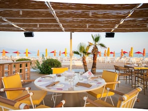 An outdoor dining area with round tables and yellow chairs, overlooking a beach with umbrellas and lounge chairs.